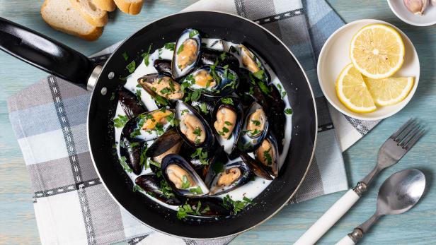 Miesmuscheln in cremiger Sauce mit Petersilie in einer Pfanne, dazu Zitronenscheiben und Baguette.