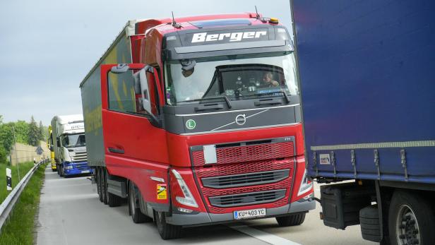 Ein roter Lkw fährt dicht neben einem blauen Lkw auf einer Autobahn, weitere Lkw sind im Hintergrund zu sehen.