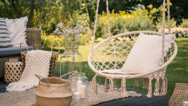 Eine gemütliche Terrasse mit Hängesessel, Pölster, Teppich, Körben, Glasvasen mit Blumen und Kerzen im Grünen.