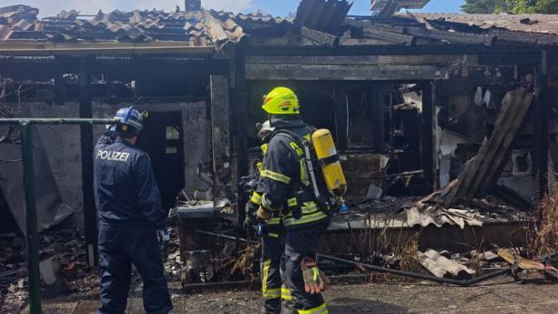 Zwei Feuerwehrleute und eine Polizistin stehen vor einem ausgebrannten Gebäude, während weitere Einsatzkräfte auf einer Drehleiter arbeiten.