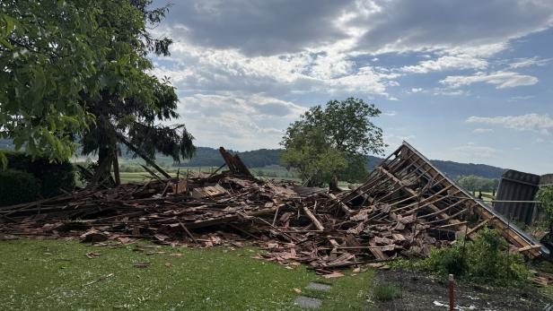 Ein eingestürztes Holzgebäude liegt in Trümmern auf einer Wiese, umgeben von Bäumen und ländlicher Landschaft.