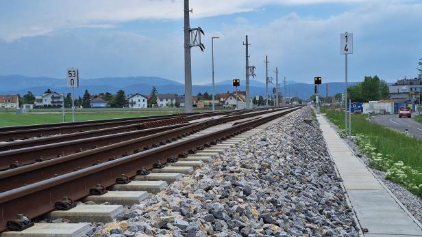 Zwei Bahngleise verlaufen parallel neben einem Gehweg, im Hintergrund sind Häuser und Berge zu sehen.