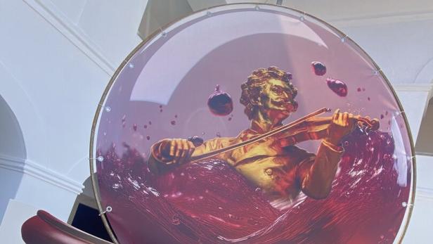 Eine goldene Figur spielt Geige in einer Glaskugel, die mit roter Flüssigkeit und Blasen gefüllt ist.
