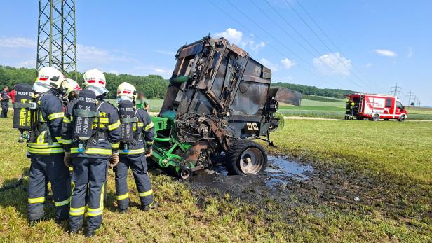 Mehrere Feuerwehrleute stehen vor einer ausgebrannten landwirtschaftlichen Maschine auf einem Feld, im Hintergrund ein Einsatzfahrzeug.