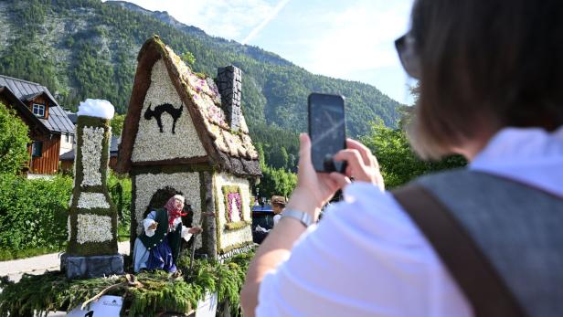 Eine Person fotografiert ein blumengeschmücktes Hexenhaus auf einem Wagen vor einer Bergkulisse.