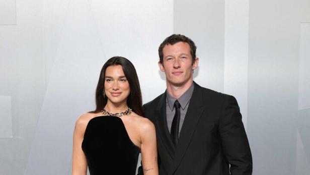 Dua Lipa in einem schwarzen Kleid mit goldenen Fransen posiert neben Callum Turner im Anzug.