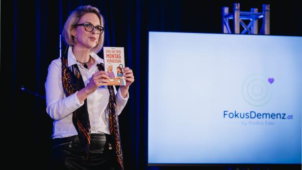 Eine Frau mit Brille hält ein Buch mit dem Titel „Oma hat den Montag vergessen“ vor einem Bildschirm mit dem Logo „FokusDemenz.at“.