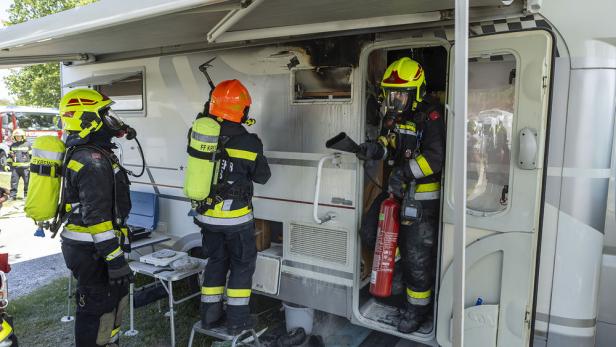 Drei Feuerwehrleute mit Atemschutz löschen einen Brand in einem Wohnmobil, das an der Tür und Wand beschädigt ist.
