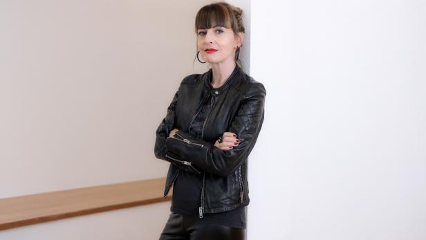 Frau mit dunkler Lederjacke und verschränkten Armen lehnt an einer weißen Wand.
