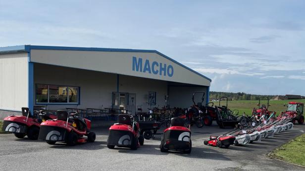 Vor einem Gebäude mit der Aufschrift „MACHO“ stehen mehrere rote Rasenmäher und landwirtschaftliche Maschinen.