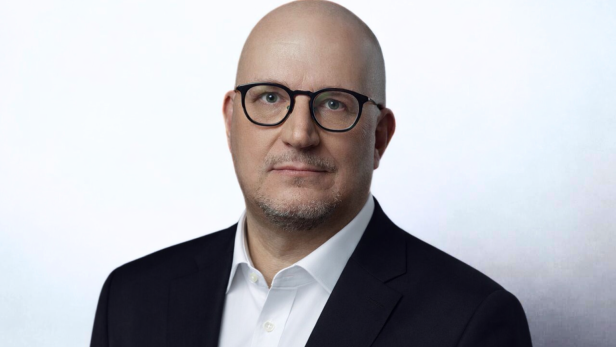 ORF-Chef-Bewerber Johannes Larcher mit Brille, Bartstoppeln und dunkles Sakko vor hellem Hintergrund.