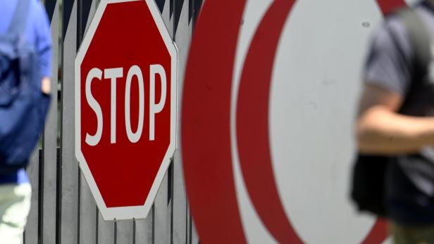 Ein rotes Stoppschild steht vor einem Zaun, daneben ein rundes Verkehrszeichen und unscharfe Personen im Vordergrund.