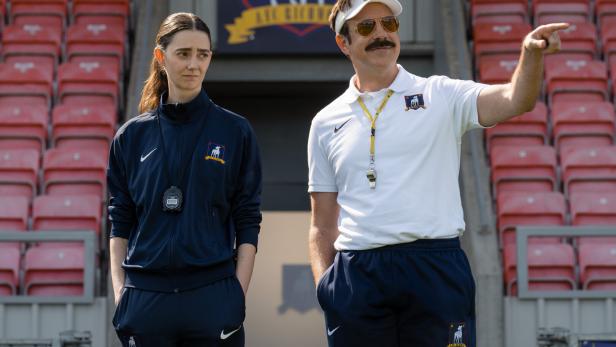 Ted Lasso zeigt auf dem Fußballfeld mit einer Frau im Trainingsanzug neben ihm.