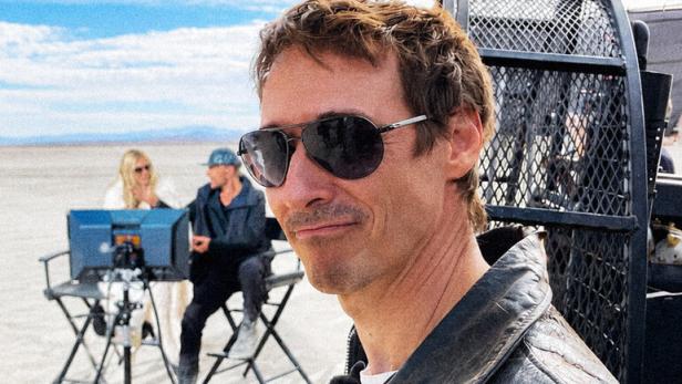 Ein Mann mit Sonnenbrille und Lederjacke steht im Vordergrund, im Hintergrund sitzen zwei Personen an Monitoren in einer Wüstenlandschaft.