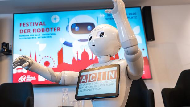 Ein humanoider Roboter mit Tablet hebt die Arme, im Hintergrund eine bunte Tafel mit dem Schriftzug „Festival der Roboter“.