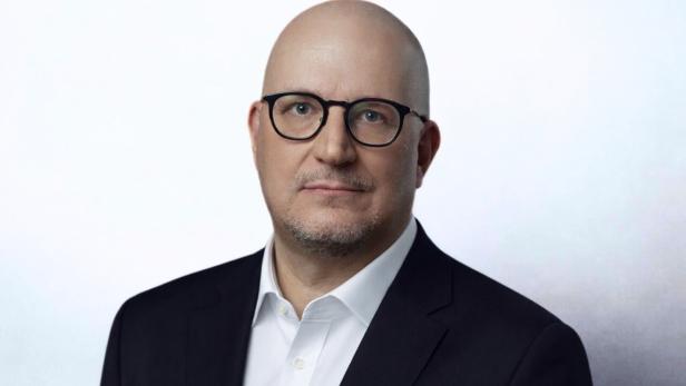 Ein Mann mit Glatze und Brille trägt ein dunkles Sakko und ein weißes Hemd vor hellem Hintergrund.