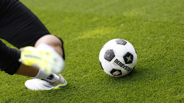 Ein Fußball liegt auf grünem Rasen, daneben ist ein Bein mit weißem Sportschuh in Bewegung zu sehen.