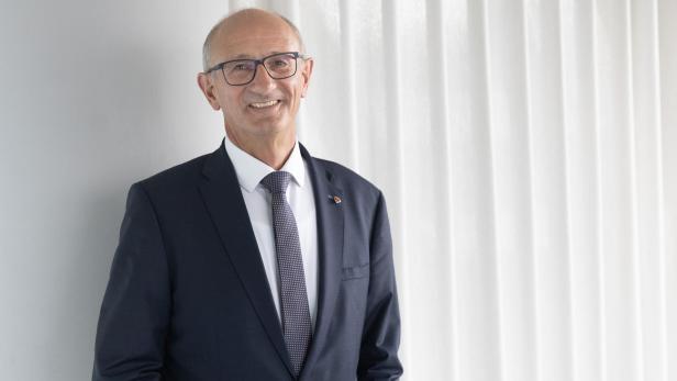 Ein älterer Mann mit Brille und Anzug steht lächelnd vor einem weißen Vorhang.