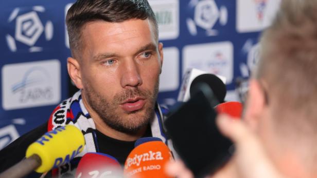 Lukas Podolski mit Schal spricht in mehrere Mikrofone vor blauem Hintergrund.