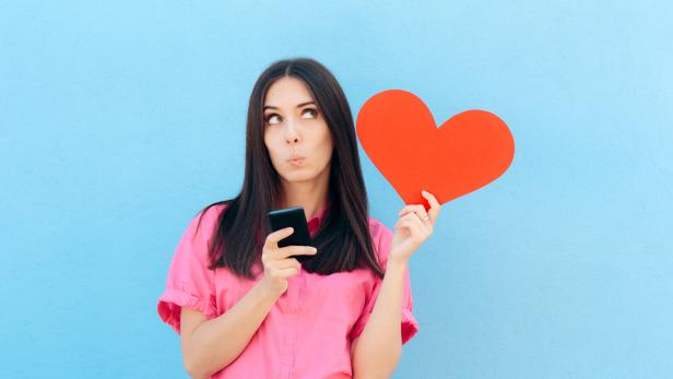 Frau in rosa Hemd hält ein rotes Herz und ein Smartphone vor blauem Hintergrund und schaut nachdenklich nach oben.