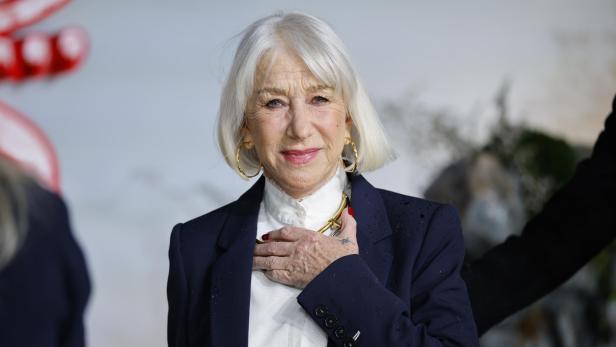 Helen Mirren lächelt in einem dunklen Blazer mit goldenen Ohrringen und einer Halskette.