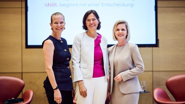 Drei Frauen stehen lächelnd nebeneinander in einem modernen Konferenzraum mit Stühlen und Präsentationsbildschirm.