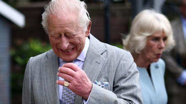 König Charles III. lächelt in einem grauen Anzug, während Camilla im Hintergrund zu sehen ist.
