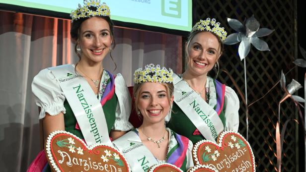 Drei junge Frauen in Tracht mit Blumenkränzen und Schärpen halten Lebkuchenherzen mit der Aufschrift „Narzissenkönigin“ und „Narzissenprinzessin“.