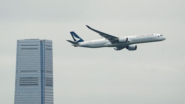 Ein Cathay Pacific Flugzeug fliegt vor einem hohen Gebäude in leicht bewölktem Himmel.