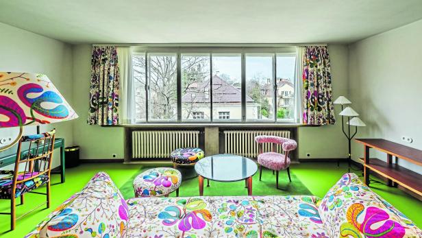 Ein Wohnzimmer mit bunten Möbeln und floralen Mustern vor einem Fenster mit Blick auf Häuser.