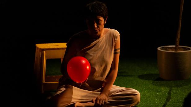 Eine Person sitzt im Schneidersitz auf grünem Boden und hält einen roten Ballon, im Hintergrund steht ein Blumentopf.