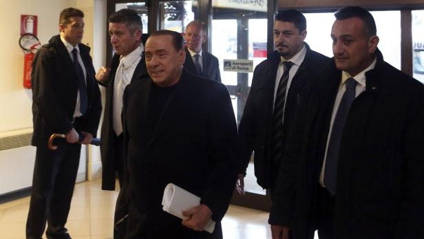 Silvio Berlusconi wird von mehreren Männern in Anzügen begleitet.