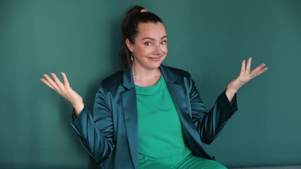 Eine Frau in grüner Kleidung und einem bläulichen Blazer sitzt mit fragenden, offenen Händen vor einem grünen Hintergrund.