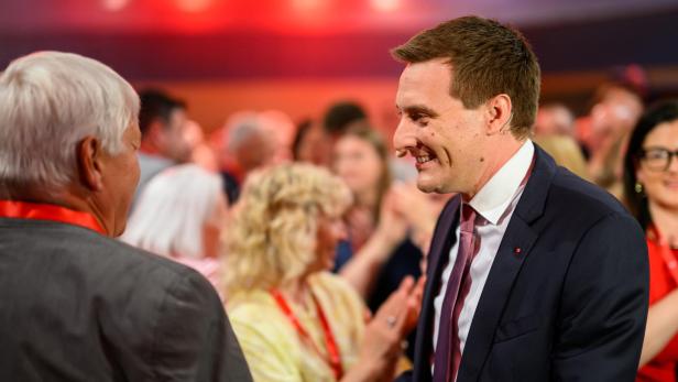 Ein Mann im Anzug lächelt und schüttelt einer anderen Person die Hand, umgeben von applaudierenden Menschen.