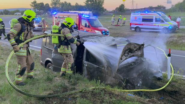 Zwei Feuerwehrleute löschen ein demoliertes Unfallfahrzeug am Straßenrand.