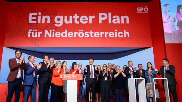 Eine Gruppe von Menschen steht auf einer Bühne vor einem großen roten Schild mit der Aufschrift „Ein guter Plan für Niederösterreich“ und applaudiert.