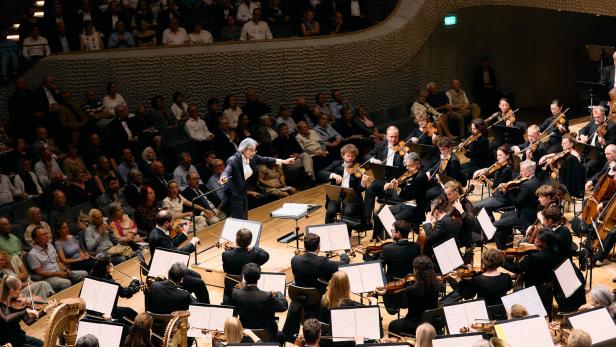 Ein Dirigent leitet ein großes Orchester auf einer Bühne vor einem aufmerksam zuhörenden Publikum.