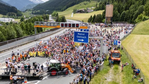Viele Menschen versammeln sich auf einer gesperrten Autobahn in den Bergen, einige halten Transparente und Schilder.