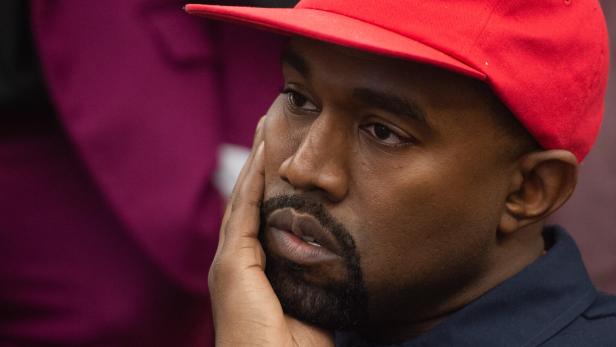 Kanye West trägt eine rote Baseballkappe und stützt nachdenklich sein Gesicht mit der Hand ab.