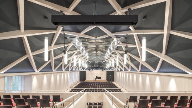 Großer moderner Konzertsaal mit geometrischer Decke, hängenden Leuchten, roten Stühlen und einem Flügel auf der Bühne.