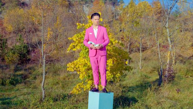 Ein Mann im pinken Anzug steht auf einem hellblauen Podest in einer herbstlichen Landschaft und hält eine Taube.