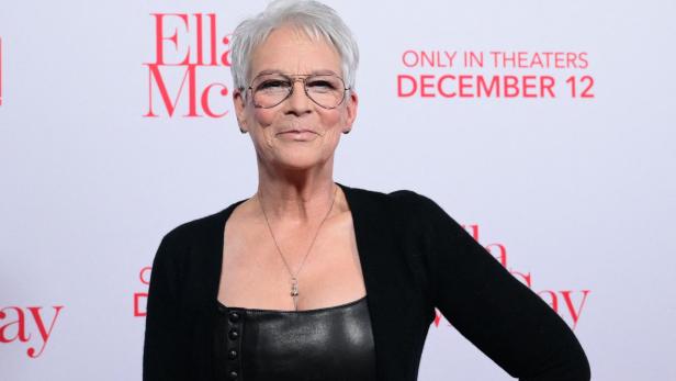 Jamie Lee Curtis trägt eine schwarze Jacke und eine Brille vor einem Werbeplakat.