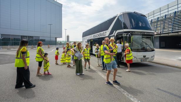 Mehrere Erwachsene und Kinder in gelben Warnwesten stehen vor einem Doppeldeckerbus mit der Aufschrift „Flughafen Wien Besucherwelt“.