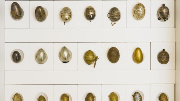Eine Sammlung von verzierten, eiförmigen Objekten ist in einem weißen Regal ausgestellt.