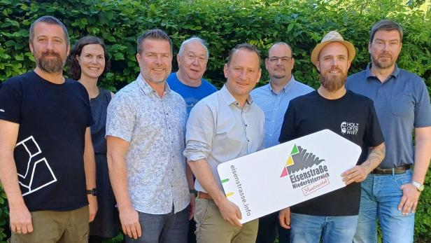 Acht Personen stehen lächelnd nebeneinander im Freien, zwei halten ein Schild mit der Aufschrift „Eisenstraße Niederösterreich“.