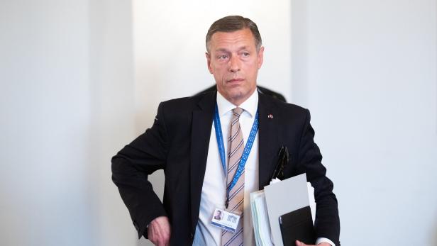 Ein Mann im Anzug mit Akten und Ausweis um den Hals steht vor hellem Hintergrund.