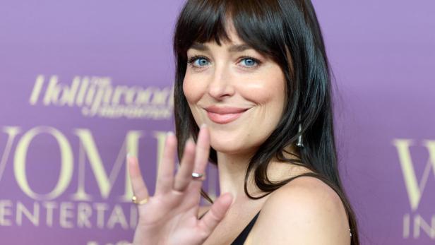 Dakota Johnson lächelt und winkt vor einem lila Hintergrund.