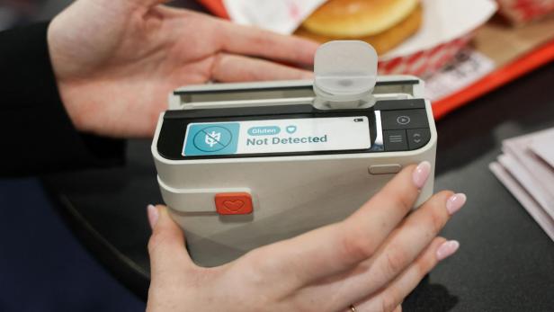 Eine Person hält ein Gerät, das anzeigt, dass in einem Lebensmittel kein Gluten nachgewiesen wurde, im Hintergrund liegt ein Burger.
