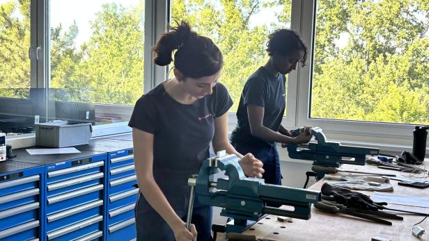 Zwei Personen arbeiten an Werkbänken mit Schraubstöcken und Werkzeugen vor großen Fenstern mit Blick ins Grüne.