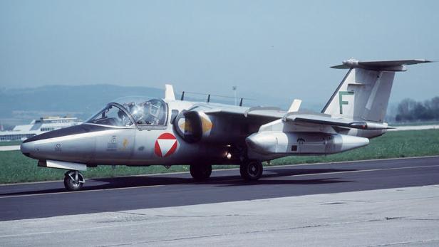Ein silbernes zweistrahliges Militärflugzeug mit rotem Emblem steht auf einer Startbahn im Freien.